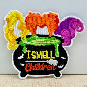 May include: Embroidered patch depicting a black cauldron with green bubbling liquid and the text "I SMELL Children" in yellow and orange. The patch includes orange, yellow, and purple hair designs, and a white border.
