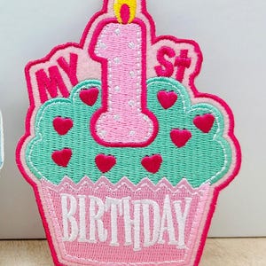 Puede incluir: Un parche bordado con forma de cupcake y una vela rosa en la parte superior. El cupcake tiene glaseado verde menta con pequeños corazones rosas, un envoltorio rosa y el texto "MY 1ST BIRTHDAY" en hilo blanco y rosa.