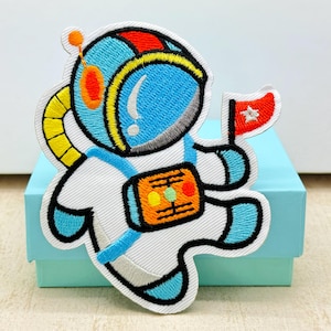 May include: Embroidered patch featuring a cartoon astronaut in a blue and white spacesuit, holding a red flag with a white star. The astronaut has a blue helmet with yellow and orange accents. The patch is on a light blue box.