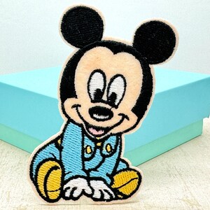 May include: An embroidered iron-on patch featuring a cartoon baby Mickey Mouse wearing a blue jumpsuit with yellow shoes. The patch is on a light blue background.