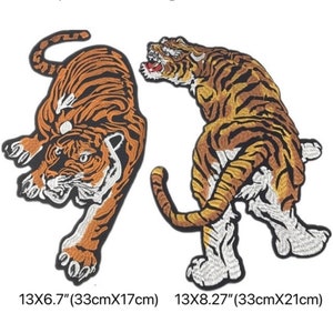 Könnte beinhalten: Zwei gestickte Bügelbilder von Tigern. Der Tiger links hockt und blickt nach links. Der Tiger rechts steht und blickt nach rechts. Beide Tiger sind orange und schwarz mit weißen Streifen. Die Patches sind 33 cm breit. Der Patch links ist 17 cm hoch und der Patch rechts ist 21 cm hoch.