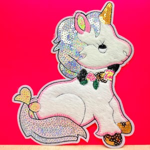 May include: A white unicorn with a pink mane and tail, decorated with iridescent sequins and a floral garland. The unicorn is sitting on a pink background.