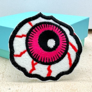May include: Embroidered eye patch featuring a white background, black outline, a bright pink iris, and red veins. This patch is a unique accessory for clothing or bags.