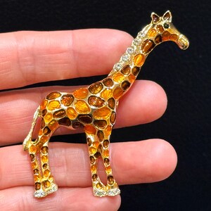 May include: A gold-tone giraffe brooch featuring a polished metal frame filled with translucent amber, orange, and dark brown enamel patches. The giraffe's hooves are accented with small, clear, sparkling rhinestones along the bottom edges.