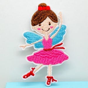 May include: Embroidered patch featuring a ballerina with brown hair, a red bow, and rosy cheeks. She wears a pink dress, a red belt, and red ballet shoes. The ballerina has blue wings and is in a dancing pose.