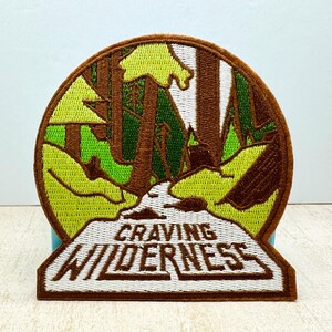 Puede incluir: Parche bordado marrón con una escena de bosque verde y marrón y el texto "CRAVING WILDERNESS" en blanco.
