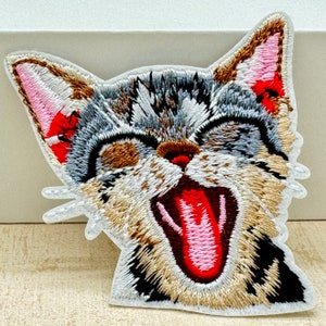 Peut inclure: Écusson brodé représentant le visage d'un chat bâillant. Le chat a une fourrure grise, marron et beige avec des oreilles intérieures roses et une bouche rouge. L'écusson est bordé de blanc.