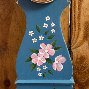 May include: A blue wooden clock with a white face and black hands. The clock is decorated with pink and white flowers and green leaves. The clock is a traditional Swedish style clock.