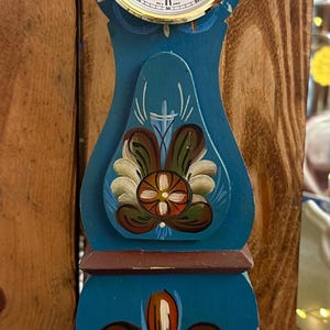 May include: A blue and brown painted wooden clock with a white face and gold trim. The clock face reads "Hermle Quartz".