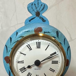 May include: A blue and gold painted clock with Roman numeral markings and a quartz movement. The clock has a decorative top with a floral design.