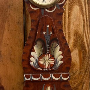 May include: A wooden clock with a painted design in red, white and blue. The clock face is white with black Roman numerals and a gold trim. The clock is on a wooden base.