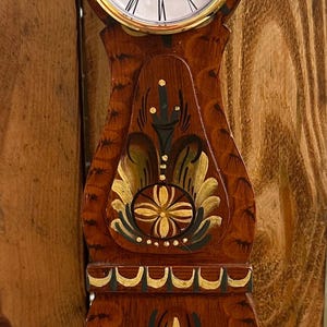 May include: A wooden clock with a painted design in the style of traditional Swedish folk art. The clock face is white with black Roman numerals and gold hands. The clock is labelled "QUARTZ".