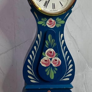 May include: A blue and white painted wooden clock with a gold rim and Roman numeral face. The clock is decorated with pink roses and green leaves. The clock is made in Germany and is quartz.