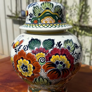 Könnte beinhalten: Eine weiße Keramikvase mit Deckel, verziert mit bunten Blumenmustern in Rot, Gelb und Orange. Der Deckel ist mit einer kleinen dekorativen Figur gekrönt. Die Vase steht auf einer Holzoberfläche.