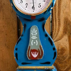 May include: A blue, decorative clock with a white clock face and gold trim. The clock has a unique shape with painted floral designs in red, yellow, and white. The clock measures approximately 10 inches tall.