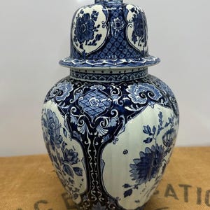 Può includere: Un vaso di zenzero in ceramica blu e bianca con coperchio, con motivi floreali e una statuina di leone sulla parte superiore. Il vaso ha un corpo bulboso e un coperchio con bordo decorativo. Il design generale è ornato e dettagliato.