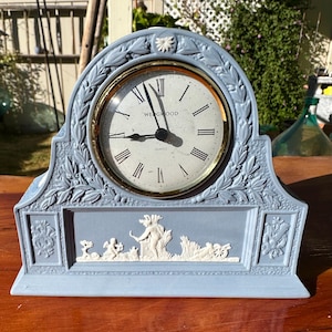 Puede incluir: Un reloj de cuarzo Wedgwood azul claro con un bisel dorado y números romanos. El reloj presenta un diseño decorativo en relieve con motivos florales y figuras clásicas. El reloj está sobre una superficie de madera.