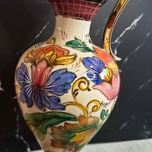 May include: A white ceramic pitcher with a gold handle and a floral design. The pitcher has a wide base and a narrow neck. The flowers are painted in shades of pink, blue, yellow, and green.