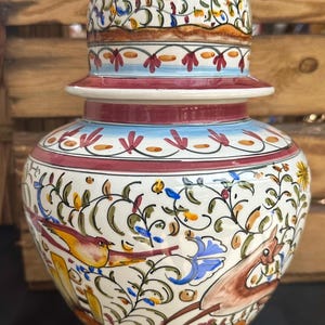 Pode incluir: Um jarro de cerâmica branca com tampa vermelha e um desenho floral com pássaros. O jarro tem um corpo redondo e uma base larga. O desenho floral é em tons de azul, verde, amarelo e vermelho.