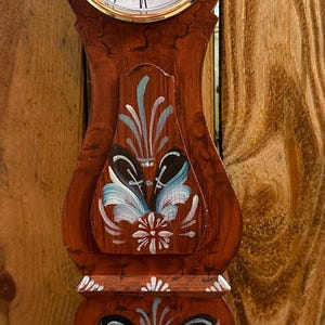 May include: A wooden clock with a painted floral design. The clock face is white with black Roman numerals and black hands. The clock is on a wooden base.