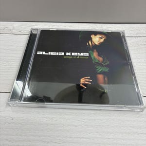 May include: A compact disc case featuring the album "songs in A minor" by Alicia Keys. The cover art displays a woman in a green and black striped top and a green hat against a dark background. The artist's name and album title are in white.
