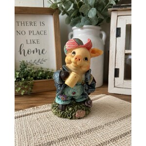 May include: A small figurine of a pig wearing a blue denim jacket, light blue overalls, and a red and white striped cap. The pig holds a small tan mug and stands on a textured green base. The figurine features detailed facial expressions.