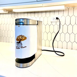 May include: A stainless steel coffee maker with a white removable cup holder. The cup holder has a cartoon illustration of a coffee cup with an umbrella and the text "Brew-Brella".