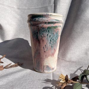 May include: A handmade ceramic tumbler with a unique glaze. The colors include pink, green, and white, creating a marbled effect. The tumbler has a cylindrical shape with a slightly textured rim. The overall aesthetic is rustic and artistic.