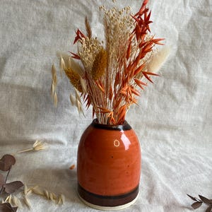 Könnte beinhalten: Eine kleine, orangefarbene Keramikvase mit braunem Rand und einem weißen Streifen am Boden. Die Vase ist mit getrockneten Blumen in Orange-, Beige- und Weißtönen gefüllt.