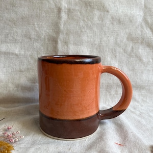 May include: A ceramic mug with a brown rim and base, and a bright orange body. The mug has a rounded handle.