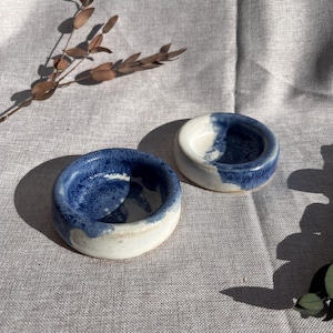 May include: Two small, handmade ceramic bowls. The bowls are round with a white base and a blue interior and rim. The blue colour has a speckled appearance. The bowls are displayed on a neutral-coloured linen surface.