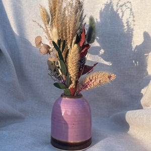 Könnte beinhalten: Ein dekoratives Arrangement aus Trockenblumen in einer kleinen, handgefertigten, lavendelfarbenen Keramikvase. Die Vase hat ein dunkelbraunes Band am Boden. Die Trockenblumen umfassen Weizen, Pampasgras und andere strukturierte Elemente.