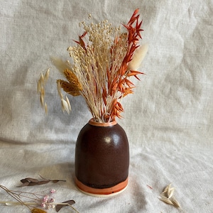 Könnte beinhalten: Eine kleine, braune Keramikvase mit einem Trockenblumenarrangement. Die Vase hat eine abgerundete Form mit einem helleren orangefarbenen Band am Boden und am Rand. Die Trockenblumen sind in Orange-, Beige- und Weißtönen gehalten.