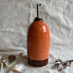May include: A ceramic olive oil bottle with a brown and orange glaze. The bottle has a metal pour spout and a brown rim.
