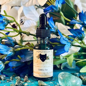 May include: A dark glass bottle of "Goddess Isis" ritual oil, with a black dropper top. The label features a black cat silhouette and the words "BrightEyed Botanicals." The background includes blue flowers, green leaves, and small stones.