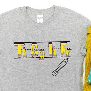 May include: Gray t-shirt with a graphic of the letters "Tt Gg Ll Ff" written in a yellow and pink pencil style font. The letters are on a lined paper background with a pencil drawn below the letters.