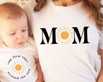Peut inclure: Un t-shirt blanc avec le mot "MOM" en lettres noires et un soleil jaune. Un bébé dans un body blanc avec le texte "my first trip around the sun" et un soleil est tenu par une personne.