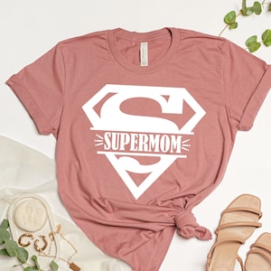 May include: A dusty rose t-shirt with a white graphic of a superhero logo and the text "SUPERMOM" inside the logo.