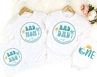 Puede incluir: Camisetas blancas y un body con diseños a juego. Las camisetas presentan las frases "RAD MOM", "RAD DAD" y "RAD BRO" en color turquesa, con un sol, una palmera y un gráfico de olas. El body dice "ONE" con un gráfico de olas y sol.