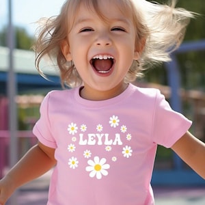 May include: A pink t-shirt with a white daisy design and the name "LEYLA" printed on the front. The shirt is being worn by a young child who is smiling and laughing.