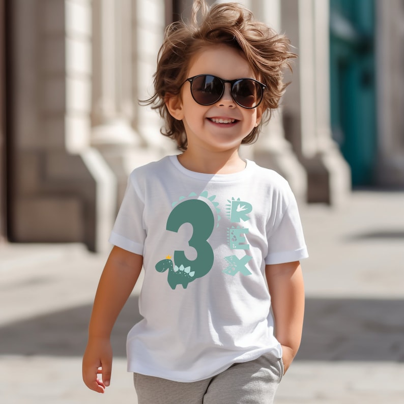 May include: A white t-shirt with a green number 3 and a dinosaur graphic. The dinosaur is green with a yellow crown and blue accents. The text "Rex" is printed in blue and white on the shirt.