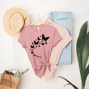 May include: A pink t-shirt with a black butterfly design. The butterflies are arranged in a curved line, with the largest butterfly at the top of the curve.