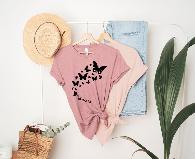 May include: A pink t-shirt with a black butterfly design. The butterflies are arranged in a curved line, with the largest butterfly at the top of the curve.