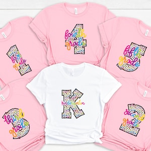 May include: Five pink t-shirts with colorful text and leopard print designs. The shirts are labeled "first grade", "second grade", "third grade", "fourth grade", and "fifth grade". One white t-shirt with a large "K" for kindergarten with leopard print and colorful text.