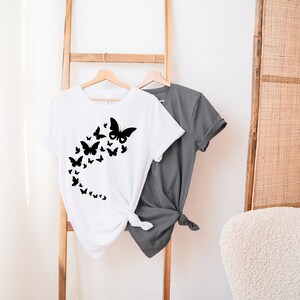 May include: A white t-shirt with a black butterfly design. The butterflies are arranged in a curved line, creating a flowing effect. The shirt is hanging on a wooden rack.