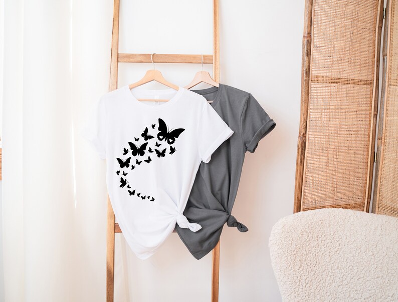 May include: A white t-shirt with a black butterfly design. The butterflies are arranged in a curved line, creating a flowing effect. The shirt is hanging on a wooden rack.