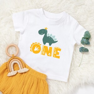 May include: White t-shirt with a green dinosaur wearing a crown and the word "ONE" in yellow letters.