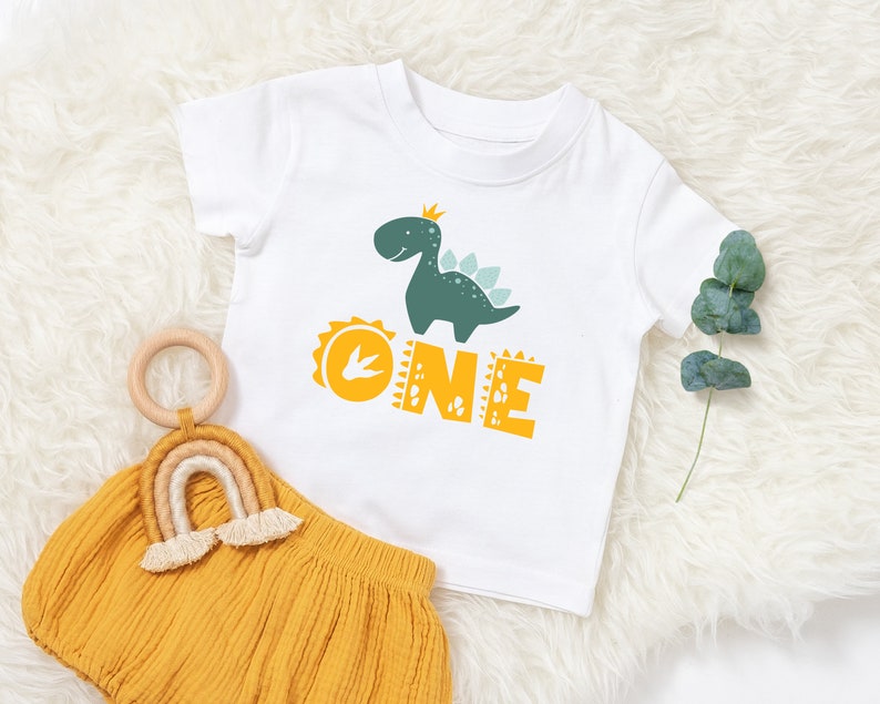 May include: White t-shirt with a green dinosaur wearing a crown and the word "ONE" in yellow letters.
