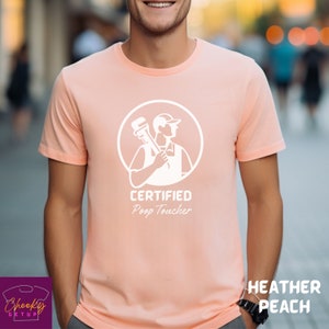 May include: A peach colored t-shirt with a white graphic of a plumber holding a wrench. The text "CERTIFIED Poop Toucher" is printed below the graphic.