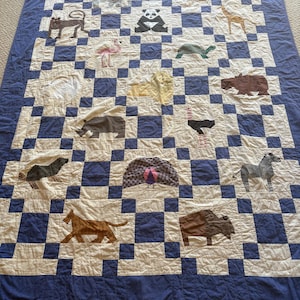 May include: A rectangular quilt featuring a blue and white checkerboard pattern with various animal appliques including a panda, giraffe, flamingo, turtle, lion, hippo, rhino, ostrich, peacock, zebra, buffalo, and other wildlife figures.